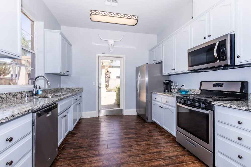Bright galley kitchen with white cabinets, stone counters, and stainless appliances.